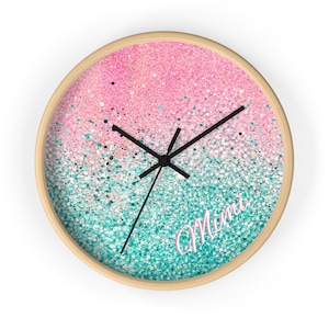 May include: Round wall clock with a light wood frame. The clock face features a gradient of pink and teal glitter with the word "Mimi" in a cursive font. Black clock hands.