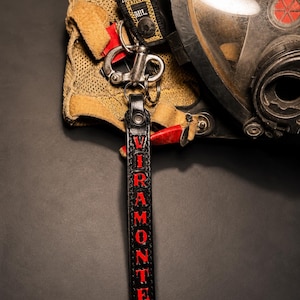 May include: A black leather keychain strap featuring the red text VIRAMONTES vertically embossed down the center. The strap is attached to a silver-toned metal swivel snap hook and a circular split ring, connected to a tan mesh fabric accessory.