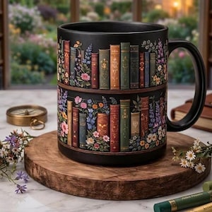 May include: A matte black ceramic mug featuring a wrap-around design of two horizontal shelves filled with colorful vintage-style books and various floral arrangements interspersed between the spines, complete with a sturdy handle on the right side.