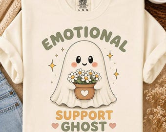 May include: A cream-colored t-shirt featuring a graphic of a white ghost holding a brown flower pot filled with white daisies. The text above reads EMOTIONAL and below reads SUPPORT GHOST in green and yellow letters, accented by small stars and hearts.