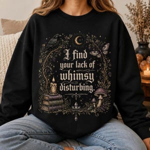 May include: A black crewneck sweatshirt featuring a graphic print with the text "I find your lack of whimsy disturbing." The design includes a raven, a crescent moon, a moth, a lit candle on a stack of three books, crystals, and mushrooms.