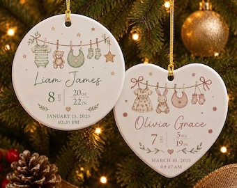 Op de afbeelding: Twee witte keramische ornamenten, één rond en één hartvormig, met illustraties van babykleding aan een waslijn. Het ronde ornament toont de naam Liam James, geboortegegevens en datum. Het hartvormige ornament toont Olivia Grace.