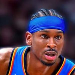 Puede incluir: Retrato de un jugador de baloncesto con una cinta azul en la cabeza y una camiseta azul y naranja. El jugador tiene una expresión concentrada, con un fondo borroso de otras personas en las gradas.
