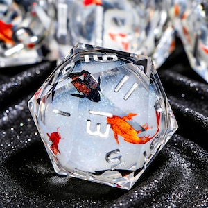 May include: A clear, multi-sided die with embedded orange and black fish. The die has silver numerals and is filled with a white, glittery substance. The die is set against a black, sparkly background.