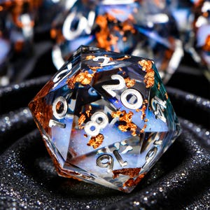 May include: A clear, multi-sided die with silver numbering. The die is filled with blue and white swirls and flecks of gold. The die is designed for role-playing games.