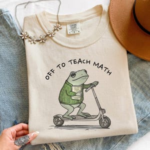 May include: A cream-coloured t-shirt with the text "OFF TO TEACH MATH." The graphic shows a frog in a green sweater, riding a scooter, and holding a book. A silver necklace with flower accents is also visible.