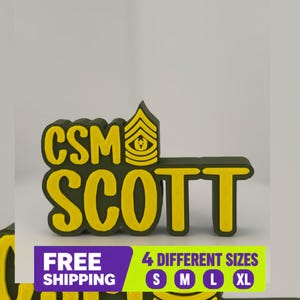 以下が含まれることがあります： 黄色い縁取りのダークグリーンの文字でCSM SCOTTと書かれた立体的な看板。文字の上には黄色い軍の階級章がある。文字の下には紫と緑のバナーがあり、FREE SHIPPINGと4 DIFFERENT SIZES S M L XLという文字が表示されている。
