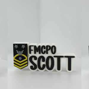 May include: A white and black nameplate with the text "FMCPO SCOTT". The nameplate is accompanied by a black, yellow, and white emblem with stars and an eagle. The nameplate is a military-themed item.