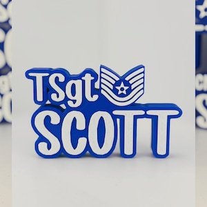 May include: A blue and white 3D-printed nameplate featuring the name "SCOTT" in large, bold letters. Above the name is "TSgt" and a military insignia with a star. The design is set against a white background.