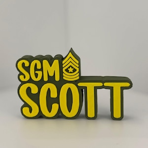 May include: A military-themed emblem in olive green and yellow. The text "SGM SCOTT" is prominently displayed, with the letters outlined in yellow. A yellow military rank insignia is also present.