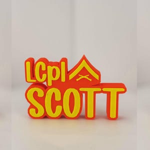 Puede incluir: Una etiqueta con el nombre de color naranja brillante con el nombre "SCOTT" en letras amarillas en negrita. La etiqueta también presenta las letras "LCpl" y una insignia militar amarilla. Este artículo probablemente está relacionado con el servicio militar.