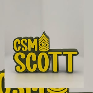 Puede incluir: Una placa con el nombre impresa en 3D con el texto "CSM SCOTT" en amarillo, delineado en verde oscuro. Encima del nombre hay una insignia de rango militar. La placa es probablemente un artículo personalizado.