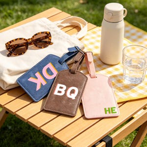 May include: Three rectangular luggage tags in blue, brown, and light pink with textured surfaces. The blue tag features the letters DK in pink and orange, the brown tag has white BQ letters, and the light pink tag displays green HE letters.
