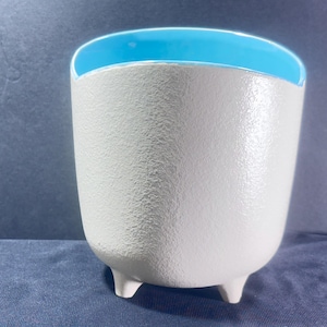 May include: A white ceramic planter with a textured surface and a bright blue interior. The planter has three small feet and a rounded shape, suitable for small plants or decorative use.