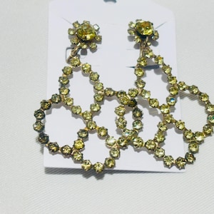 May include: A pair of chandelier earrings featuring a cluster of light green, faceted rhinestones set in a metallic frame. Each earring consists of a circular top stud connected to a larger, openwork geometric structure made of linked stones.