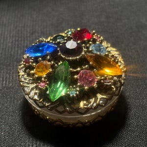 May include: A small, round, vintage trinket box with a clear base and a gold-colored filigree top. The top is adorned with an array of colorful, faceted gemstones in shades of red, blue, green, yellow, and pink.