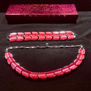 May include: A red necklace and bracelet set with a matching red box. The necklace and bracelet feature a series of connected, rectangular links. The box is a deep pink with a glittery finish. The jewelry is displayed on a black surface.