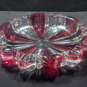 May include: A clear glass ashtray with a red rim and accents. The ashtray has a circular shape with a scalloped edge and a starburst design radiating from the center. The glass is transparent, allowing light to pass through and reflect.
