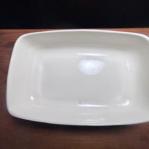 May include: A rectangular, off-white ceramic baking dish with rounded corners. The dish is empty and sits on a dark brown wooden surface. The dish has a glossy finish and is clean.