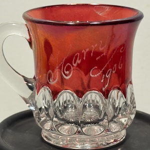 May include: A small red glass mug with a clear glass handle. The upper portion of the mug features a deep red finish, while the lower base is clear with a pressed, scalloped pattern. The text "Harry 1906" is etched into the red glass surface.