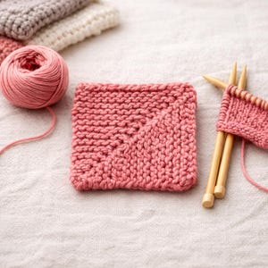 Può includere: Un quadrato di tessuto a maglia color rosa antico, con un motivo diagonale, è posto accanto a una matassa di filato abbinato e ferri da maglia in legno. Ulteriori pezzi a maglia bianchi e grigi sono sullo sfondo.