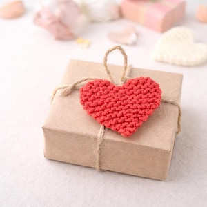 Może przedstawiać: Małe, kwadratowe pudełko prezentowe owinięte w brązowy papier i związane sznurkiem. Czerwone, dziergane serce jest przymocowane do góry pudełka. Inne dekoracyjne serca są w tle.