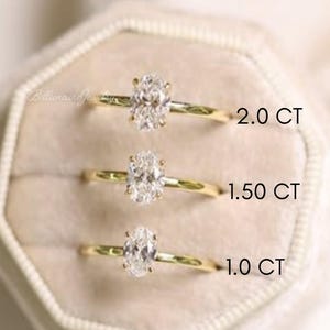 May include: Three gold engagement rings with oval-cut diamonds are displayed in a beige velvet ring box. The rings are labeled 1.0 CT, 1.50 CT, and 2.0 CT. The rings are set against a soft, neutral background.