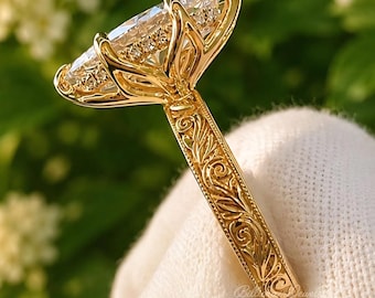 May include: A yellow gold ring featuring a large marquise-cut center stone held by a prong setting. The band is decorated with intricate, raised floral and scroll engravings that extend along the sides, showcasing detailed metalwork craftsmanship.
