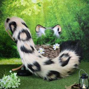 Puede incluir: Un conjunto de cola y orejas de piel sintética. La cola es de color crema con manchas negras y una punta negra. Las orejas son blancas con detalles marrones y negros. El conjunto está diseñado para parecerse a un animal manchado.