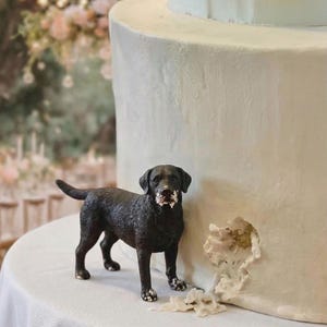 May include: A black Labrador retriever figurine with white frosting on its face and paws stands next to a white cake with a bite taken out of it. The cake is on a white tablecloth.