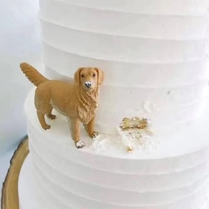 Può includere: Figurina di cane golden retriever marrone chiaro su una torta glassata bianca. Il cane è dettagliato e si trova sullo strato superiore della torta. La torta ha un design strutturato e scanalato. La torta è decorata con foglie d'oro.
