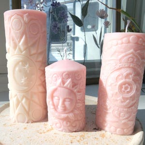 May include: Three decorative pillar candles in varying sizes. The tallest candle is off-white with a sun, moon, and star design. The medium candle is pink with a sun face design. The third candle is pink with a floral pattern.
