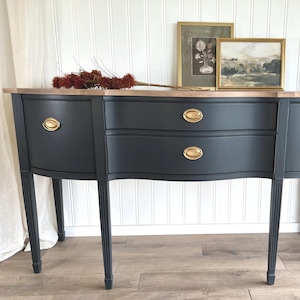 May include: A dark gray buffet with a light wood top and gold hardware. The buffet has two drawers and curved sides. Two framed pictures and a dried floral arrangement are on top of the buffet.