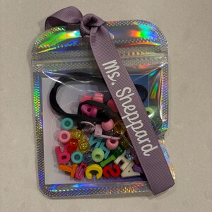 Op de afbeelding: Een doorzichtige, holografische zak gevuld met kleurrijke kralen, letters en cijfers. Een zwarte haarband en een paars lint met de tekst "Ms. Sheppard" zijn ook inbegrepen. De zak is afgesloten met een paars lint.