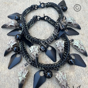 May include: A layered chain necklace featuring black metal links, multiple off-white bird skull charms with dark beaks, matte black teardrop-shaped metal pendants, and small glossy black spherical beads attached throughout the chain structure.