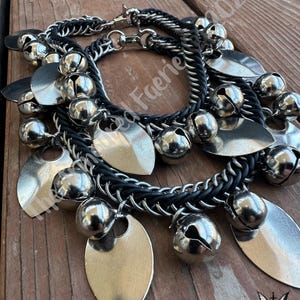 May include: A layered bracelet set featuring silver-toned metal charms and bells. The bracelets have a black, braided band and silver chain accents. Leaf-shaped charms and round bells dangle from the bracelets. The background is a wooden surface.