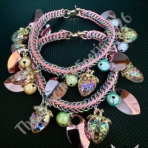 May include: Two pink chainmail bracelets with gold clasps and charms. Charms include iridescent strawberry pendants, pink leaf-shaped charms, small bells, and green and pearl-colored beads. The bracelets are on a dark green background.