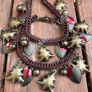 May include: A handcrafted necklace with a brown and red chainmail design. The necklace features bronze-colored Christmas tree charms, red and green leaf-shaped charms, and small bronze bells. The necklace is displayed on a wooden surface.