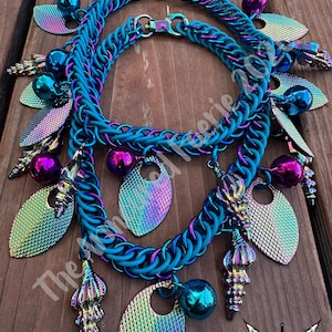 May include: Two turquoise chainmail necklaces with iridescent charms. The necklaces feature various charms, including teardrop shapes, spheres, and shell-like pendants. The image is set against a wooden background.