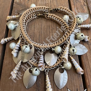 May include: A handcrafted necklace featuring a woven tan band and a chain link design. The necklace is adorned with silver-toned leaf-shaped charms, small bells, and seashell pendants. The text "The Armoury Faerie" is visible.