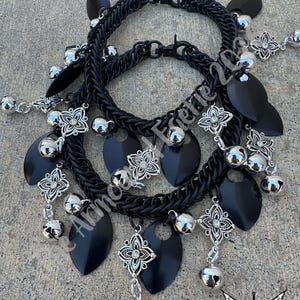 May include: Two black chainmail necklaces with silver-colored accents. Each necklace features black teardrop-shaped pendants, silver floral charms, and small silver bells. The necklaces are displayed on a textured gray surface.