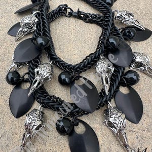 May include: A black chainmail necklace and bracelet set. The jewelry features silver-colored bird skull charms, black oval charms, and black spherical beads. The chainmail is made of small, interlocking rings.