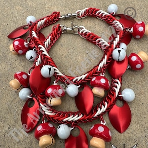 May include: A chainmail necklace featuring red and white interlocking metal rings, accented with dangling red mushroom charms with white spots, solid red teardrop-shaped metal scales, and small white jingle bells attached with silver lobster clasps.