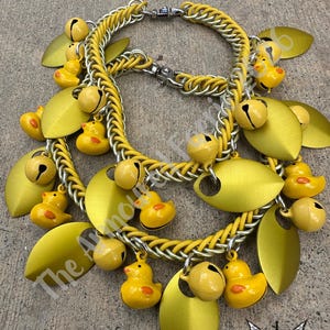May include: Two yellow and silver chain necklaces with duck and bell charms. The necklaces have yellow chainmaille and silver chain links. The charms include small yellow rubber duckies, yellow bells, and yellow leaf-shaped pendants.