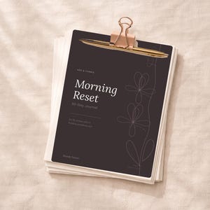 Peut inclure: Un ensemble de papier blanc avec un journal de couverture gris foncé intitulé "Morning Reset 30-Day Journal" avec une pince dorée. Le journal a un motif floral et le texte "For the woman who is building something real."