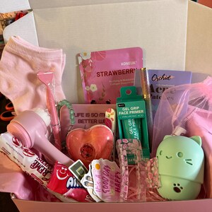May include: A pink gift box filled with beauty and self-care items. Includes a pink face brush, face masks, stickers, a heart-shaped bath bomb, candy, and a green cat-shaped item. The box is lined with pink tissue paper.