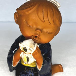 Może przedstawiać: Ceramiczna figurka dziecka w granatowym kimono, trzymającego mały biały przedmiot. Dziecko ma brązową skórę i włosy, z białą kokardą we włosach. Figurka ma błyszczące wykończenie i klęczy.