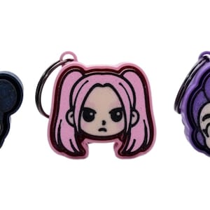 May include: Three cartoon character keyrings. The keyrings feature a black, pink, and purple colour scheme. Each keyring has a different hairstyle and facial expression. The keyrings are attached to a black metal ring.