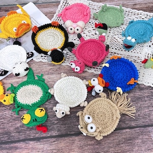 Op de afbeelding: Een verzameling kleurrijke, ronde, gehaakte onderzetters met dierenthema's, waaronder een gele eend, een zwart-witte panda, een groene kikker, een roze varken, een blauw wezen en een beige schaap met een staart van franjes.