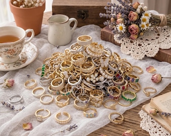 May include: A large collection of assorted rings in gold and silver tones, featuring various designs including pearl accents, colourful gemstones, clear crystals, and intricate metalwork, piled together on a white fabric runner on a wooden table.